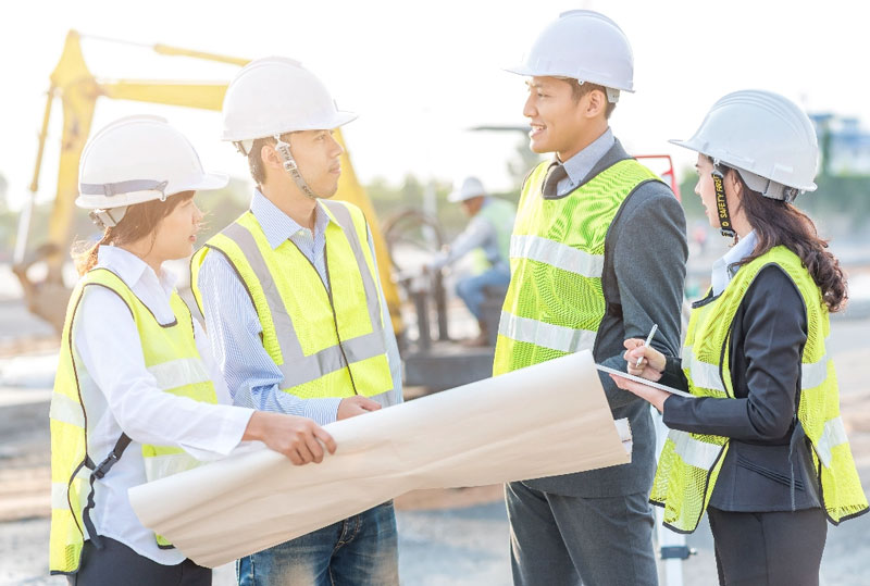 Four engineers with blueprints planning with heavy machines in the background